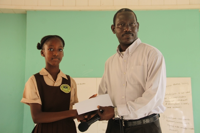 Ms. Alexzandra Weekes former student of the Joycelyn Liburd Primary School, receives her winning cheque at the local level in the Junior Category of the 2017 Florida Caribbean Cruise Association Foundation Essay Competition from Mr. John Hanley, Acting Permanent Secretary in the Ministry of Tourism on behalf of Hon. Mark Brantley, Premier of Nevis and Minister of Tourism at morning devotions at the Gingerland Secondary School on January 22, 2018