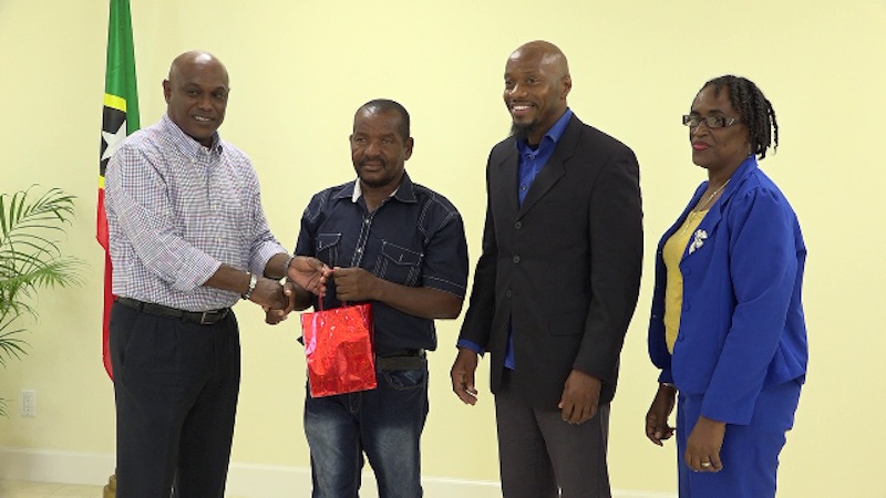 (L-r) Mr. Vernel Powell, Assistant Director of the St. Christopher and Nevis Social Security presents a token of appreciation to Mr. William “Bully” Perkins a 13-year driver and messenger with the Ministry of Health, at the Board’s Nevis Branch office at Pinney’s Estate on May 18, 2018. Looking on are Board members Mr. Timothy Caines and Mrs. Jacqueline Brooks-Jeffers look on