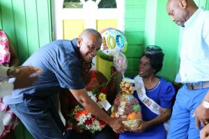 Hon. Eric Evelyn, Minister responsible for seniors on Nevis presents a gift basket in collaboration with City Drug Store as a token of appreciation to Ms. Eliza “Liza Liburd Jeffers” Flanders in celebration of her 100th birthday at her home in Hamilton on November 30, 2019, while her son Mr. Elton Liburd (right) looks on